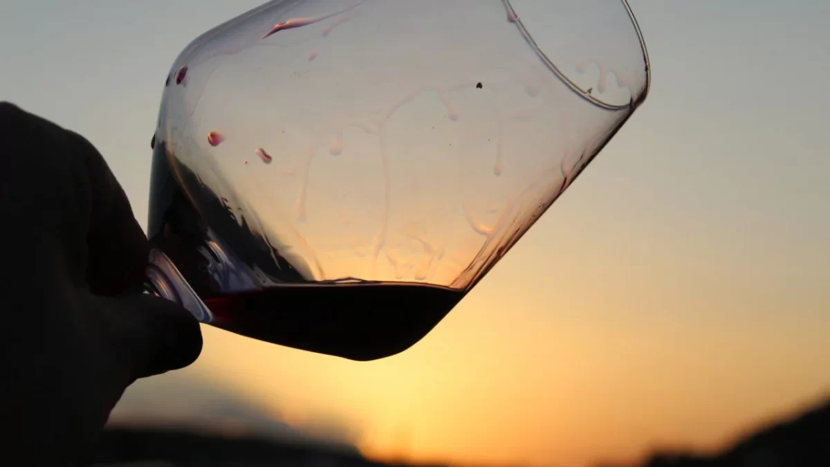 Mano sosteniendo una copa inclinada de vino tinto con un atardecer de fondo, mostrando el paisaje sereno de la Comunidad Valenciana