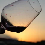 Mano sosteniendo una copa inclinada de vino tinto con un atardecer de fondo, mostrando el paisaje sereno de la Comunidad Valenciana