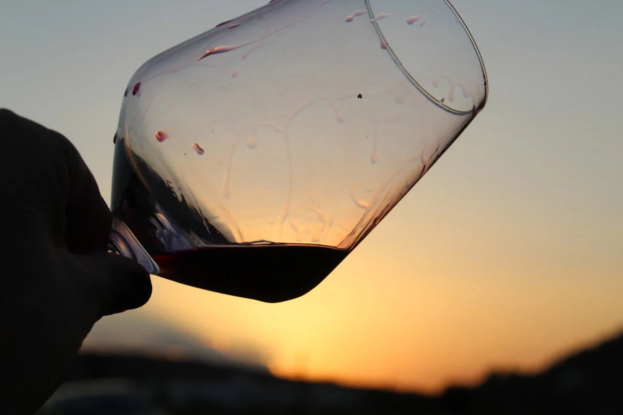 Mano sosteniendo una copa inclinada de vino tinto con un atardecer de fondo, mostrando el paisaje sereno de la Comunidad Valenciana